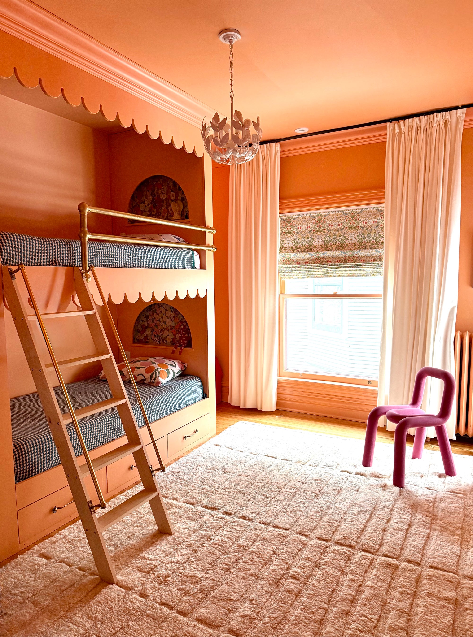 color drenched peach colored kids bedroom with floral, white chandelier. White fluffy carpet and a modern purple chair accent the room. The stylish kids bunk beds have nooks for special items and checkered kids bedsheets. 